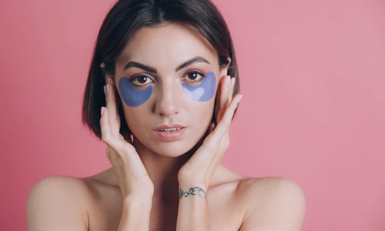 Žena s modrými gelními maskami pod očima na růžovém pozadí.