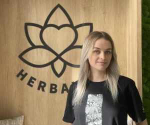 Žena stojí před dřevěnou stěnou s logem značky Herba.