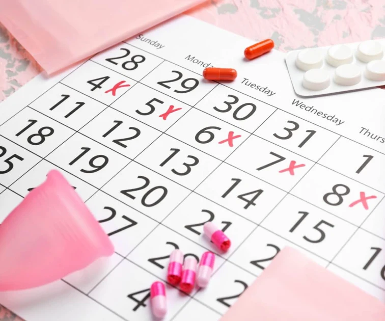 Kalendář s označením menstruace a hygienické produkty na růžovém pozadí.