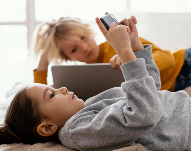 Dvě děti leží na posteli s tabletu a mobilním telefonem.