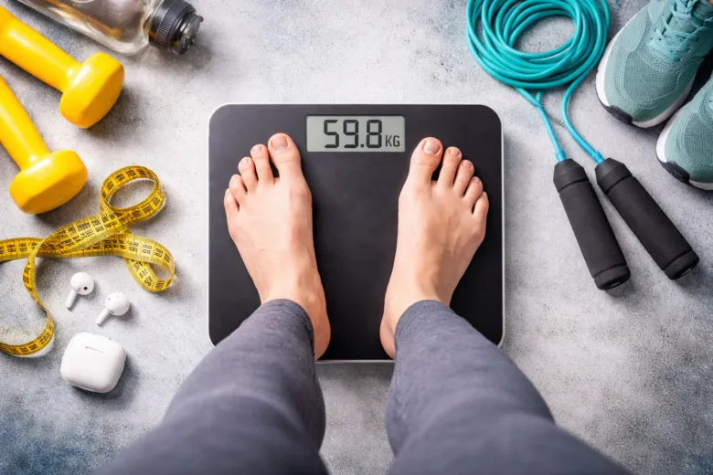 Člověk váží na vahách s fitness vybavením kolem.
