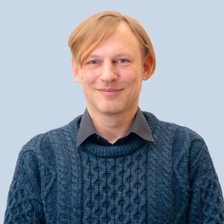 Muž v modrém svetrku s úsměvem na neutrálním pozadí.