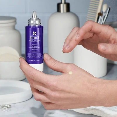Ruce aplikují krém z fialové lahvičky značky Kiehl's na kůži.