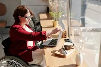 Žena v invalidním vozíku pracuje na notebooku u okna.