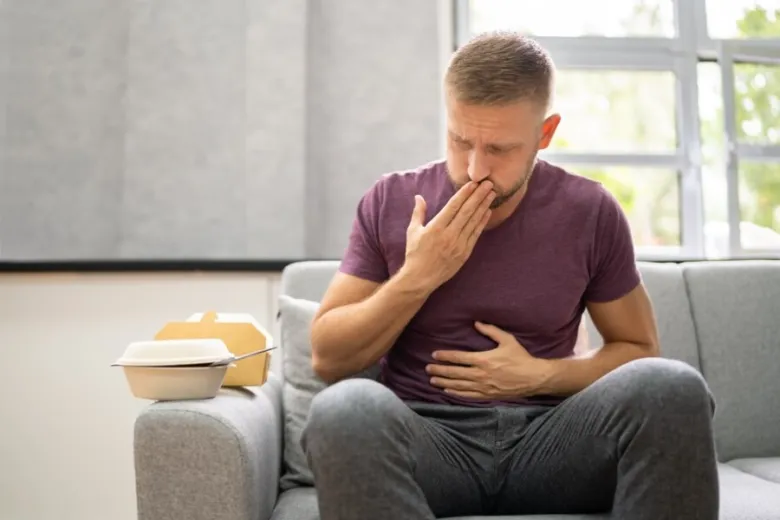 Muž sedí na gauči s bolestí břicha po jídle, typické pro reflux.