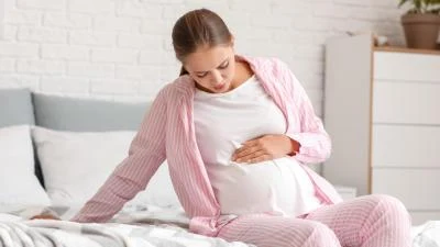 Těhotná žena sedí na posteli a drží si břicho.