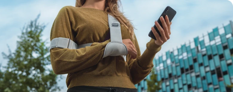 Žena s klíční kostí v ortopedickém obvazu drží mobilní telefon.
