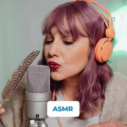 Žena s fialovými vlasy, sluchátky a peřím u mikrofonu pro ASMR.