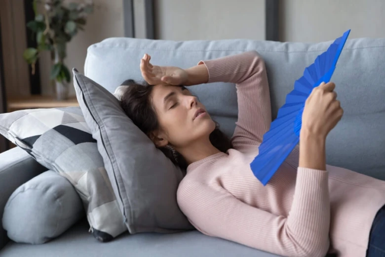 Žena leží na gauči s ventilátorem kvůli přehřívání.