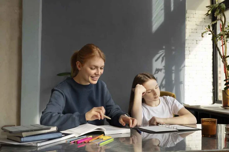 Žena pomáhá dívce studovat u stolu s učebnicemi.
